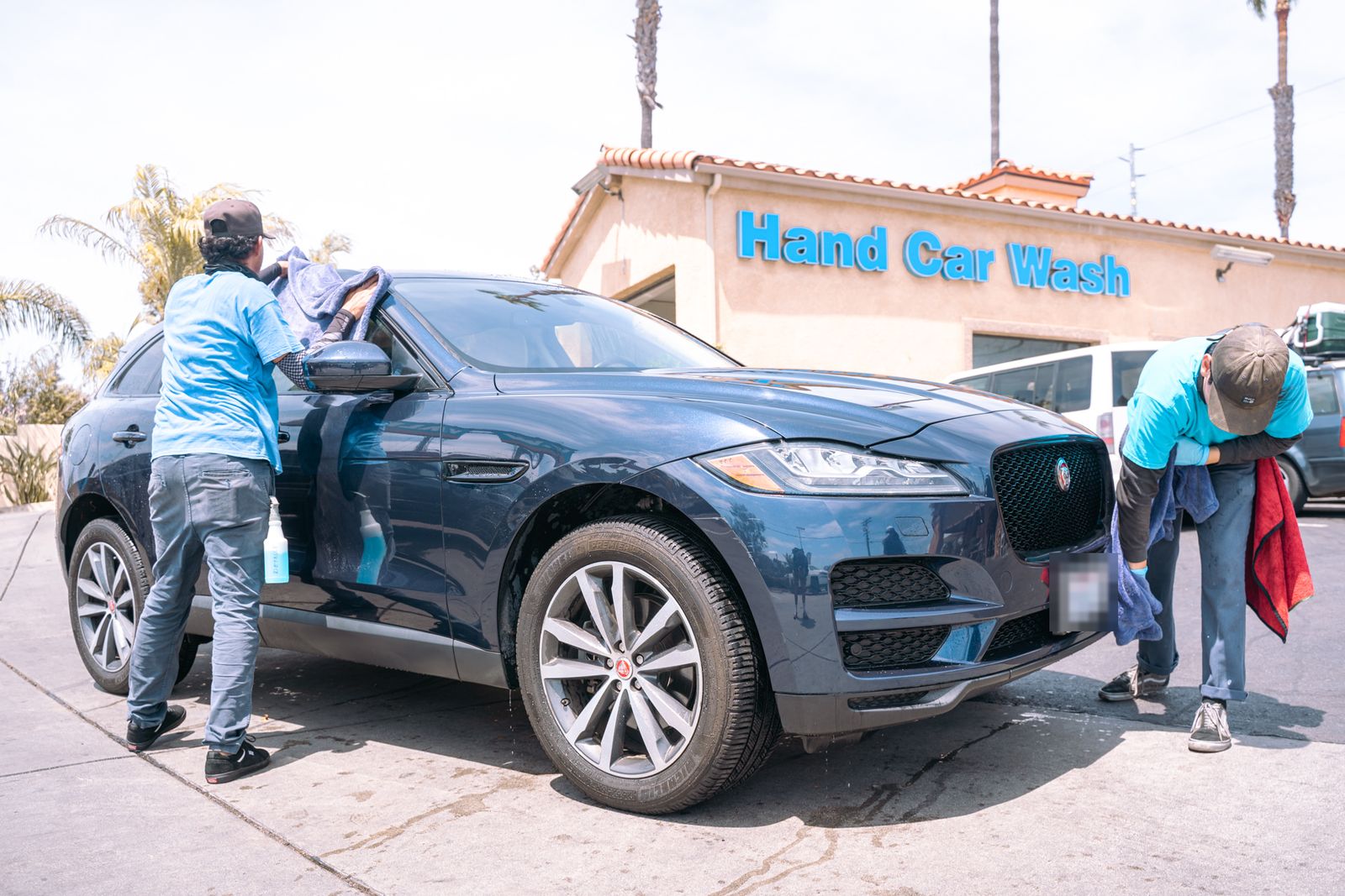 Two crew members hand-drying a navy Jaguar F-Pace in front of the Hand Car Wash building